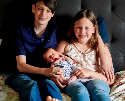 siblings hold baby on bed
